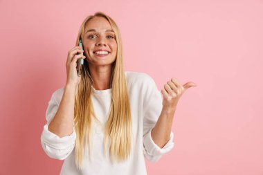 Neşeli güzel bir kız pembe arka planda izole edilmiş bir şekilde parmak işareti yapıyor ve cep telefonuyla konuşuyor.