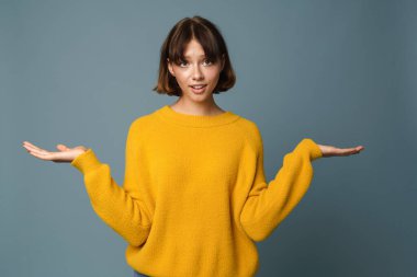 Mavi arkaplan üzerinde izole edilmiş referans gösteren kafası karışmış kadın, kopya boşluğunda işaret parmağı