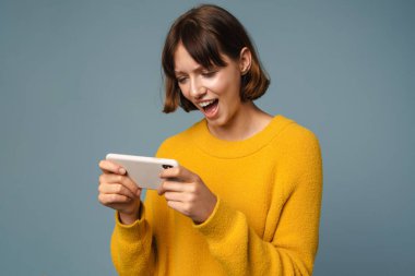 Mavi arka planda izole bir şekilde cep telefonuyla oyun oynayan mutlu çekici kız.