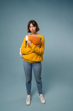 Talim kitaplarıyla poz veren mutsuz, güzel bir öğrenci kadın. Mavi arka planda, tam boy.