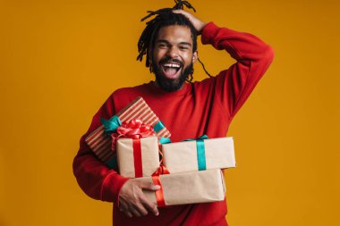 Mutlu Afrikalı Amerikalı adam, sarı arka planda izole edilmiş Noel hediyeleriyle poz verirken gülüyor.