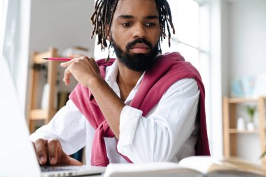 Ofiste otururken dizüstü bilgisayar ve kitapla çalışan odaklanmış Afro-Amerikan adam.