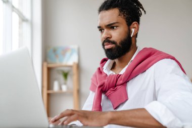 Ciddi Afro-Amerikalı kulaklıklı adam ofiste otururken dizüstü bilgisayarla çalışıyor.