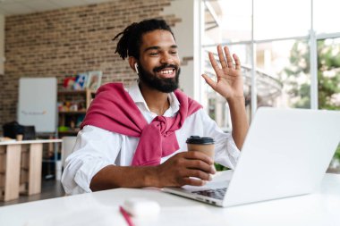 Gülümseyen Afrikalı Amerikalı adam ofiste dizüstü bilgisayarla çalışırken el sallıyor ve kahve içiyor.