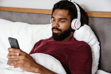 Genç Afro-Amerikalı adam yatakta müzikten hoşlanıyor, akıllı telefon kullanıyor.
