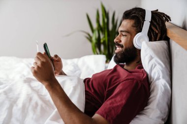 Genç mutlu Afro-Amerikalı adam yatakta müzik dinliyor, akıllı telefon kullanıyor.