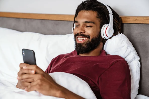 自宅でベッドに横たわっている間 携帯電話を使用してヘッドフォンで陽気なアフリカ系アメリカ人男性 — ストック写真
