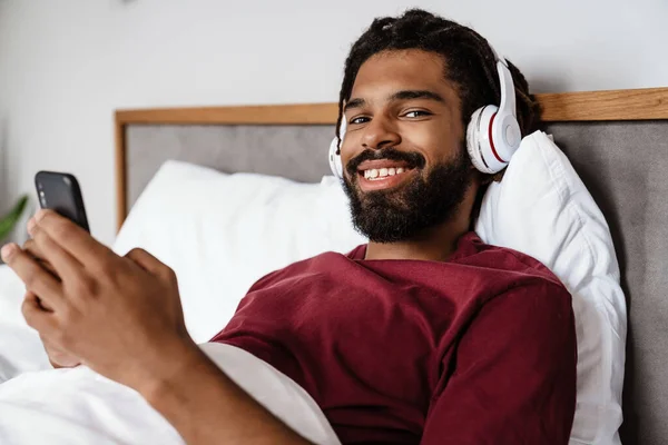 自宅でベッドに横たわっている間 携帯電話を使用してヘッドフォンで陽気なアフリカ系アメリカ人男性 — ストック写真