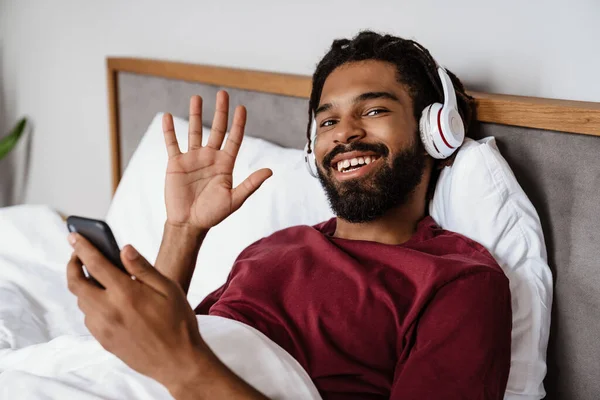 自宅で携帯電話を使用している間に手を振ってヘッドフォンで陽気なアフリカ系アメリカ人男性 — ストック写真