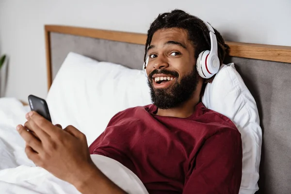 ベッドで家に横たわっている間 携帯電話を使用してヘッドフォンで興奮したアフリカ系アメリカ人男性 — ストック写真