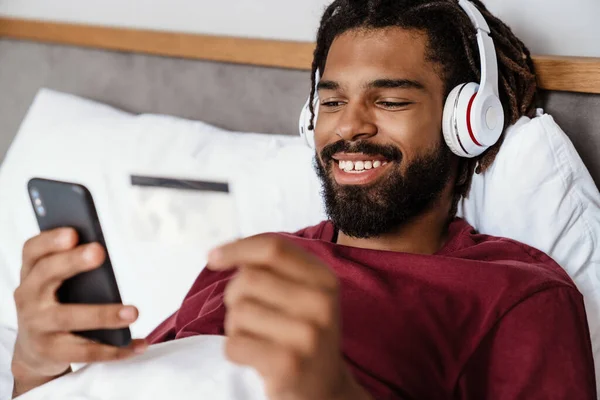 若い幸せなアフリカ系アメリカ人男性は ベッドで音楽を楽しんで スマートフォンを使用して — ストック写真