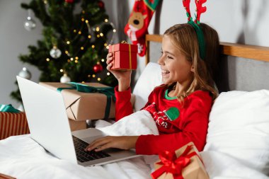 Oyuncak geyik boynuzu takan, elinde hediye kutusu olan ve evde yatarken dizüstü bilgisayar kullanan mutlu bir kız.