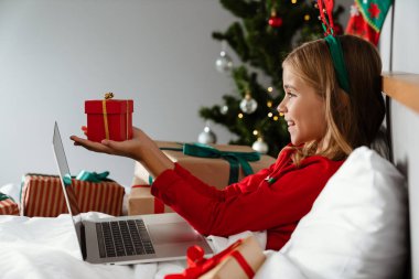 Oyuncak geyik boynuzu takan, elinde hediye kutusu olan ve evde yatarken dizüstü bilgisayar kullanan mutlu bir kız.