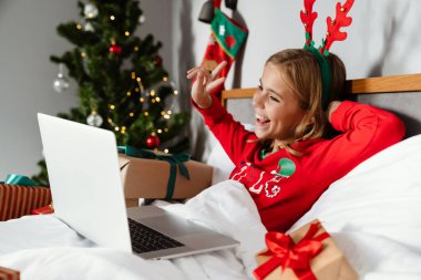 Mutlu güzel kız oyuncak geyik boynuzları takıyor barış işareti yapıyor ve evde yatarken dizüstü bilgisayar kullanıyor.