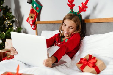 Oyuncak geyik boynuzları takan, gülümseyen ve evde yatarken dizüstü bilgisayar kullanan neşeli bir kız.