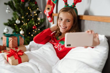 Oyuncak geyik boynuzları takan neşeli beyaz kız yatakta yatarken cep telefonundan selfie çekiyor.