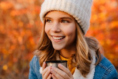 Sonbahar parkında oturmuş çay içen neşeli genç bir kız resmi.