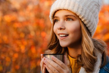 Genç, mutlu, pozitif bir kızın sonbahar parkında oturup çay içtiği görüntüsü.