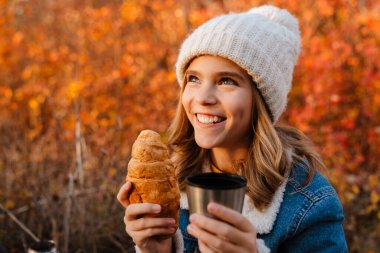 Sonbahar parkında çay içip kruvasan yiyen genç mutlu bir kız resmi.