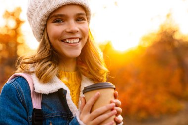 Sonbahar parkında oturup kahve içen neşeli genç bir kız resmi.