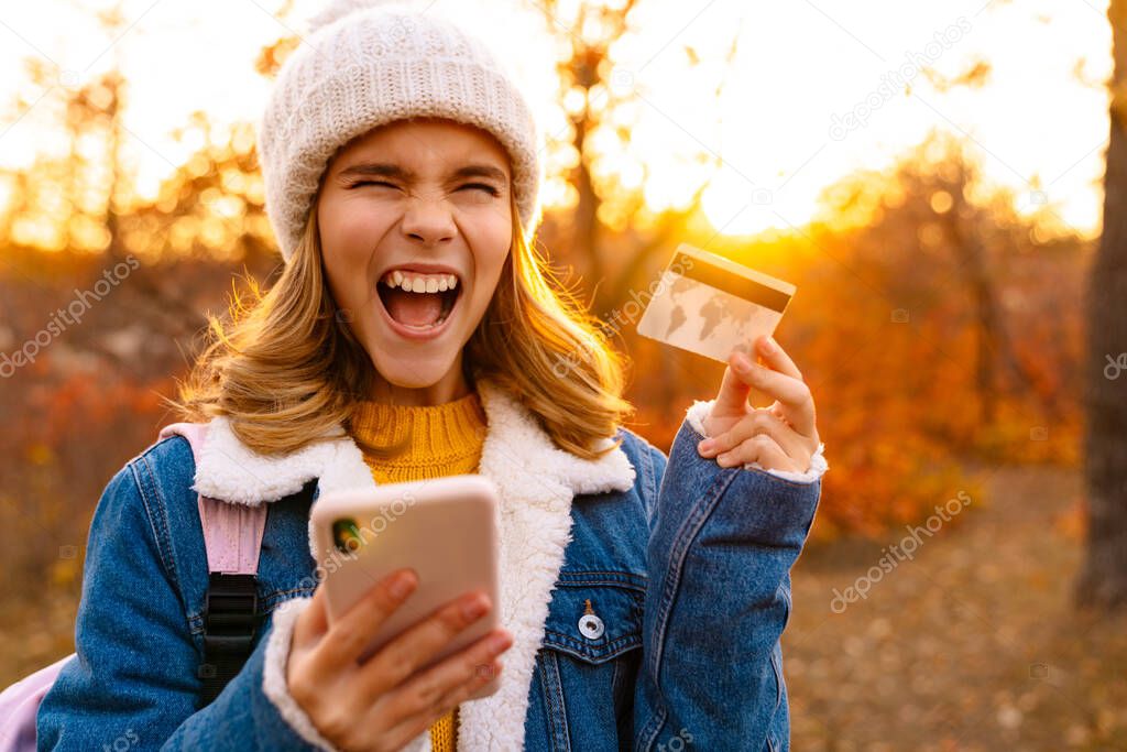 Smiling little girl showing card and mobile phone while standing in the autumn park, celebratinf