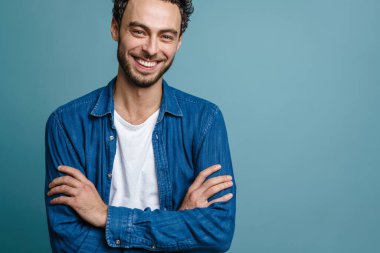 Gülümseyen ve elleri çapraz pozlar veren neşeli yakışıklı adam mavi arka planda izole edilmiş.