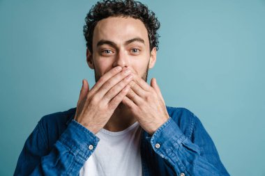 Neşeli yakışıklı adam poz veriyor ve mavi arka planda ağzını kapatıyor.