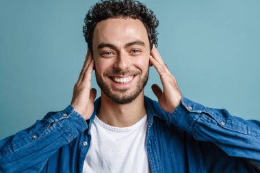 Gülümseyen ve kulaklarını kapatan neşeli yakışıklı adam mavi arka planda izole edilmiş.