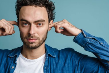 Beyaz tenli yakışıklı adam mavi arka planda poz veriyor ve kulaklarını tıkıyor.