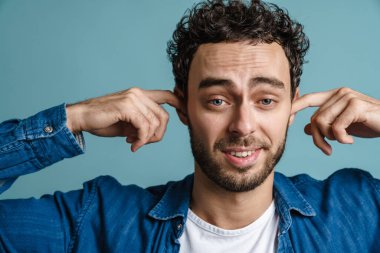 Kafası karışmış yakışıklı adam mavi arka planda poz verip kulaklarını tıkıyor.