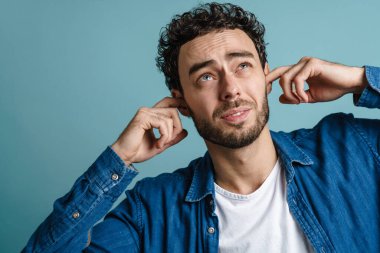 Beyaz mutsuz adam mavi arka planda poz veriyor ve kulaklarını tıkıyor.