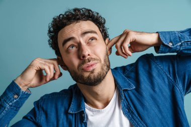 Beyaz mutsuz adam mavi arka planda poz veriyor ve kulaklarını tıkıyor.