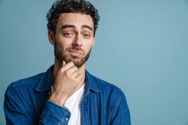 Kafası karışmış yakışıklı adam poz veriyor ve mavi arka planda izole edilmiş kameraya bakıyor.