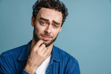 Kafası karışmış yakışıklı adam poz veriyor ve mavi arka planda izole edilmiş kameraya bakıyor.