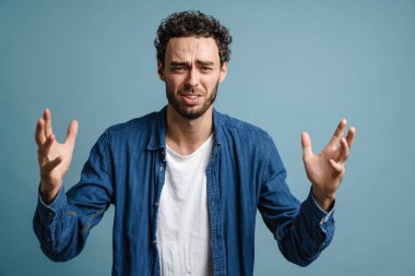 Mutsuz, yakışıklı, elleri yukarıda poz veren adam mavi arka planda izole edilmiş.