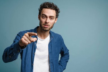Memnun olmayan yakışıklı adam mavi arka planda izole edilmiş kameraya bakıyor ve işaret ediyor.