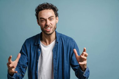 Mavi arka planda, elleri yukarıda kameraya poz veren öfkeli yakışıklı adam.