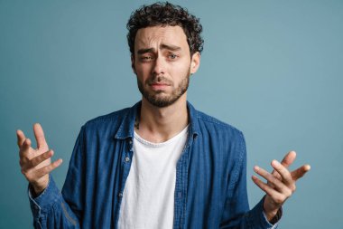 Kafası karışmış yakışıklı adam ellerini kaldırıp mavi arka planda yalnız başına kameraya poz veriyor.
