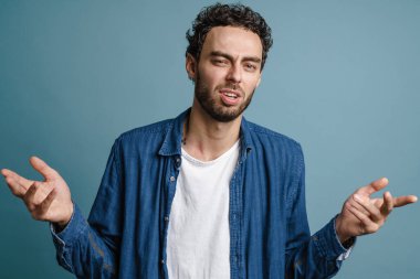 Kafası karışmış yakışıklı adam ellerini kaldırıp mavi arka planda yalnız başına kameraya poz veriyor.