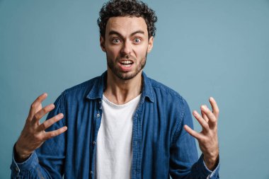 Mavi arka planda, elleri yukarıda kameraya poz veren öfkeli yakışıklı adam.