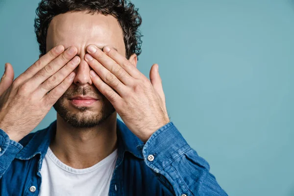 Caucasian handsome guy posing and covering his eyes isolated over blue background