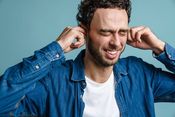 Cheerful handsome guy with eyes closed plugging his ears isolated over blue background