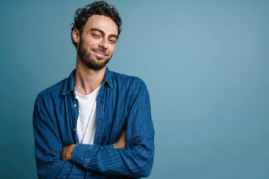 Gülümseyen ve elleri çapraz pozlar veren neşeli yakışıklı adam mavi arka planda izole edilmiş.