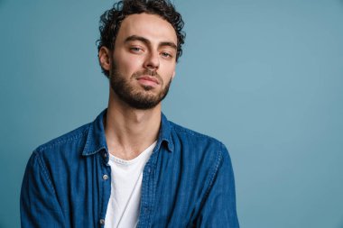 Tıraşsız yakışıklı adam poz veriyor ve mavi arka planda izole edilmiş kameraya bakıyor.
