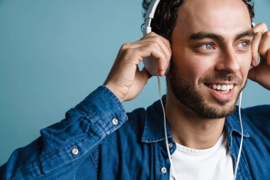 Gülümseyen yakışıklı adam mavi arka planda kulaklıkla müzik dinliyor.