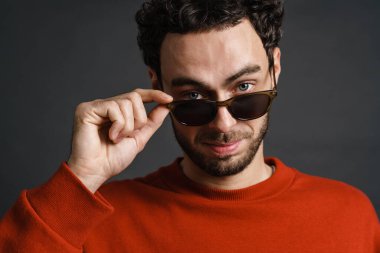 Tıraşsız, güneş gözlüklü yakışıklı adam gri arka planda yalnız başına poz veriyor ve kameraya bakıyor.