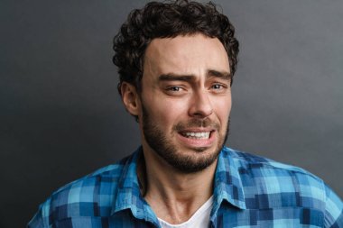 Tıraşsız, korkmuş, poz veren ve gri arka planda izole edilmiş kameraya bakan adam.