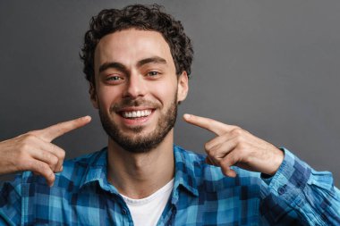 Neşeli yakışıklı adam gülümsüyor ve parmaklarını yüzüne doğrultuyor. Gri arka planda soyutlanmış.