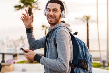 Kulaklıklı gülümseyen adam el sallıyor ve gezinti alanında yürürken cep telefonu kullanıyor.