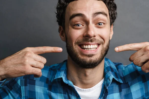 Joyful handsome guy smiling and pointing fingers at his face isolated over grey background